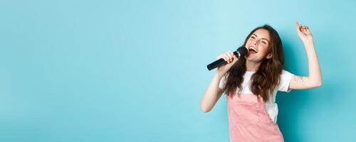 Happy young woman perform song, singer holding microphone, dancing and singing at karaoke, standing over blue background photo