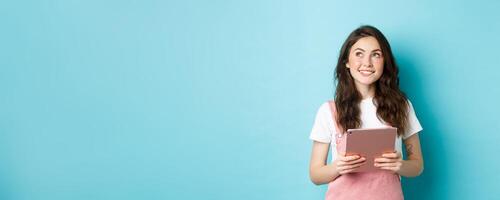 Beautiful dreamy girl smiling, holding tablet in hands and looking at upper left corner logo with thoughtful face, standing over blue background photo