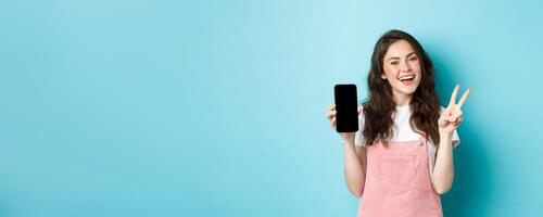 Cute young woman smiling and showing v-sign with empty smartphone screen, demonstrate app or mobile store, standing against blue background photo