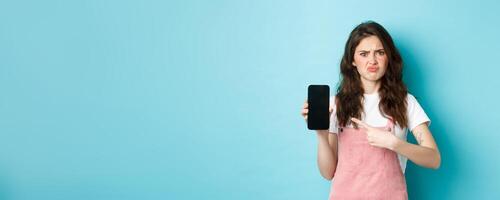 Skeptical and upset young woman frowning, sulking disappointed while pointing at empty smartphone screen with bad online offer or app, standing against blue background photo