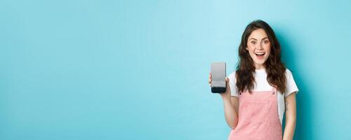 Image of beautiful young woman look surprised and excited, showing empty smartphone screen, your logo or app on mobile display, standing over blue background photo
