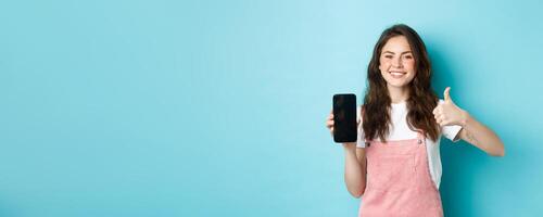This is good. Smiling cheerful woman showng empty smartphone screen and thumb up to recommend app or online store, standing against blue background photo