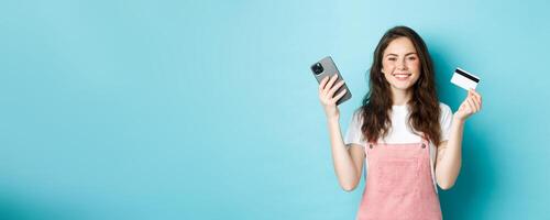 Smiling cute girl showing plastic credit card and smartphone, order something in store via mobile app, shop online, or pay for order, standing against blue background photo