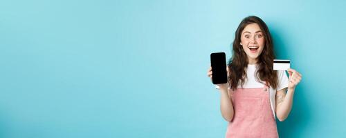 Look here. Excited young cute girl showing empty mobile screen and plastic credit card, look amazed at camera, standing over blue background photo