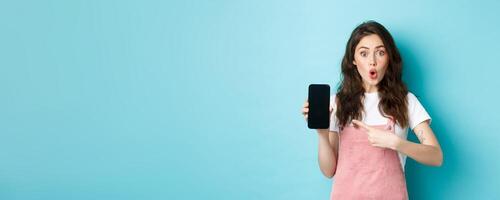 Impressed young modern woman pointing at smartphone screen, say wow and stare amazed at camera, showing awesome application, new online store, standing over blue background photo