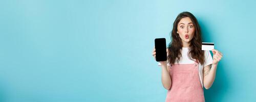 Portrait of young beautiful woman showing plastic credit card and empty smartphone screen, look amused and interested, showing interesting app, standing over blue background photo