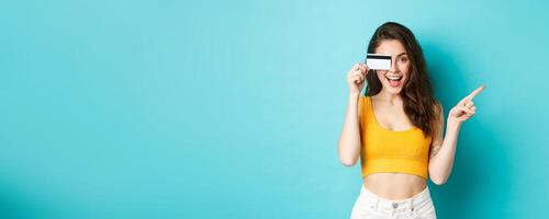 Look there. Stylish modern woman showing plastic credit card, smiling and pointing right, showing way to banner or logo, standing against blue background photo