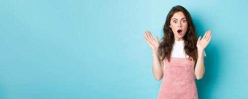Portrait of shocked attractive woman gasping, spread hands sideways and stare startled at camera, hear worrying terrible news, looking with disbelief, standing against bue background photo