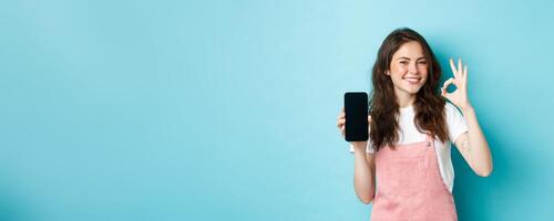 Very good. Smiling young woman showing product online on empty smartphone screen, recommending your brand, make ok sign, standing satisfied against blue background photo