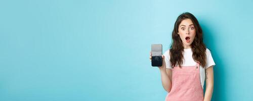 Shocked beautiful girl drop jaw and show empty smartphone screen, demonstrate your app or online store on blank phone display, standing against blue background photo