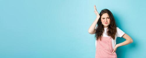 Image of troubled young girlfriend holding hand on head and frowning, remember or forget something important, standing bothered against blue background photo