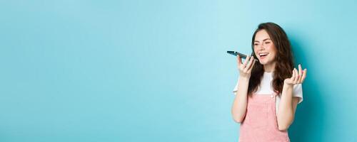 Portrait of smiling woman holding phone near lips and talking, using app translator on smartphone or recording voice message, speaking with speakerphone, standing over blue background photo