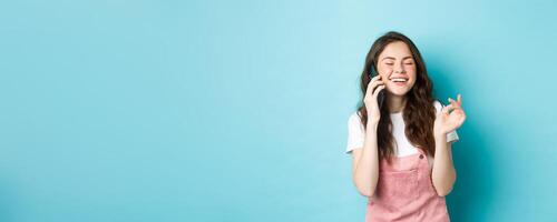 retrato de linda y elegante niña riendo mientras chateando en teléfono, participación teléfono inteligente cerca oído y sonriente feliz, teniendo casual conversación, en pie terminado azul antecedentes foto