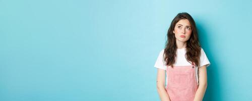 Portrait of sad and gloomy young brunette girl with glamour make up, looking at upper left corner upset and thoughtful, feeling moody, standing against blue background photo