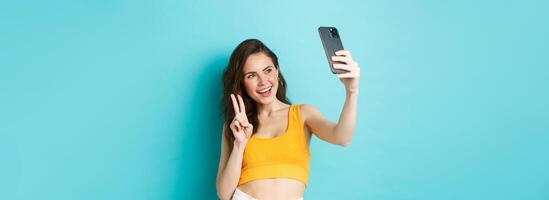 Technology and lifestyle concept. Stylish young woman taking selfie in her summer clothes, showing v-sign at smartphone camera, standing over blue background photo