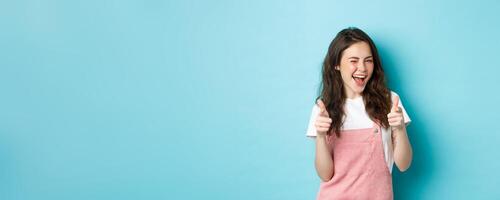 You can do this. Beautiful cheerful brunette girl winking and smiling, pointing fingers at camera, praising good job, inviting to event, standing against blue background photo