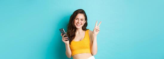 Technology and lifestyle concept. Beautiful cheerful girl sending positive vibes, smiling and showing peace sign, using cellphone, standing against blue background photo