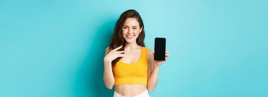 Stylish caucasian woman in summer outfit showing empty smartphone screen and smiling, demonstrate an app or online store, standing over blue background photo