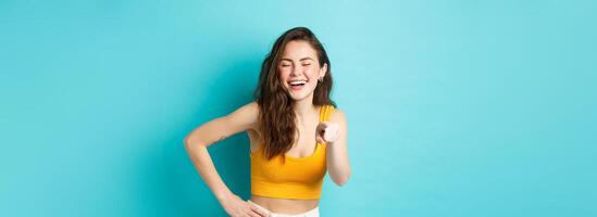 Young cheerful woman with beautiful smile, laughing at something funny, pointing finger at camera, chuckle and having fun, standing over blue background photo