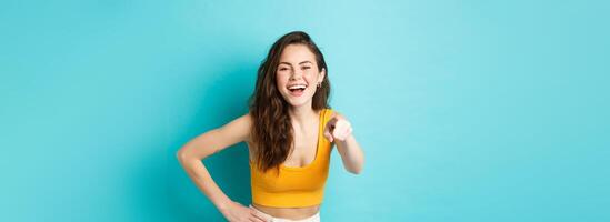 Young attractive woman with perfect body in summer clothes, laughing and smiling, pointing finger at camera, choosing you, standing over blue background photo