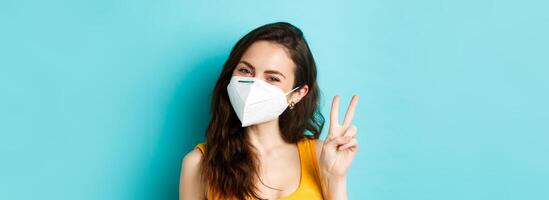 Covid-19, coronavirus and social distancing. Young positive woman in respirator showing v-sign, standing against blue background photo