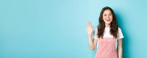 Hello nice to meet you. Beautiful friendly girl waving hand to say hi, looking happy and smiling while greeting you, giving warm welcome, standing over blue background photo