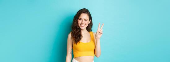 Summer and lifestyle concept. Beautiful girl tourist in crop top, posing with v-sign and smiling happy at camera, standing against blue background photo