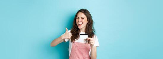 Satisfied beautiful modern girl showing plastic credit card and thumb up, recommend bank or store, standing over blue background photo