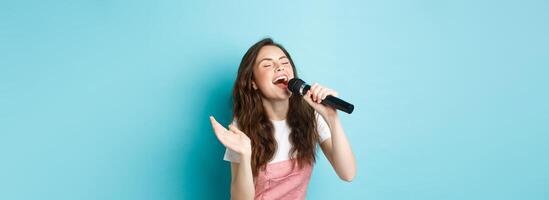 Carefree beautiful woman perform song, singing in microphone with passion, playing karaoke, standing over blue background photo