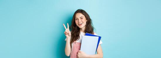 Beautiful female student showing v-sign and smiling happy, holding notebooks with study material, attending courses, standing over blue background photo