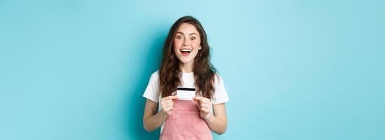 Excited beautiful female model showing plastic credit card, staring dreamy at camera, going shopping, standing over blue background photo