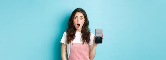 Shocked beautiful girl drop jaw and show empty smartphone screen, demonstrate your app or online store on blank phone display, standing against blue background photo