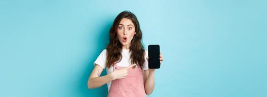 Impressed young modern woman pointing at smartphone screen, say wow and stare amazed at camera, showing awesome application, new online store, standing over blue background photo