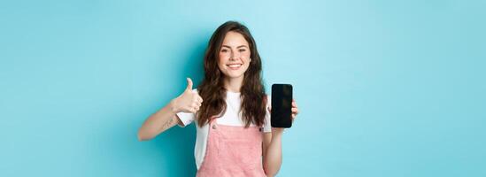 This is good. Smiling cheerful woman showng empty smartphone screen and thumb up to recommend app or online store, standing against blue background photo