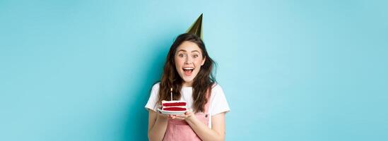 Holidays and celebration. Cheerful birthday girl in party hat holding bday cake and smiling, making wish on lit candle, standing against blue background photo