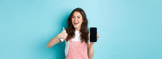 Yes this is good. Smiling cute girl showing thumb up and empty smartphone screen, recommending application or online store, standing over blue background photo