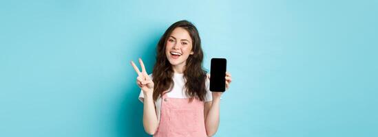 Cute young woman smiling and showing v-sign with empty smartphone screen, demonstrate app or mobile store, standing against blue background photo