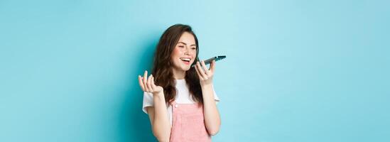 Portrait of smiling woman holding phone near lips and talking, using app translator on smartphone or recording voice message, speaking with speakerphone, standing over blue background photo