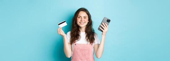 Smiling cute girl showing plastic credit card and smartphone, order something in store via mobile app, shop online, or pay for order, standing against blue background photo