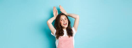 Beautiful girl mimic Easter bunny, showing rabbit ears with hands on head and smiling lovely, dancing happy, standing over blue background photo