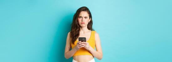 Confused and freak out young woman frowning, grimacing after reading strange message on smartphone, standing over blue background photo