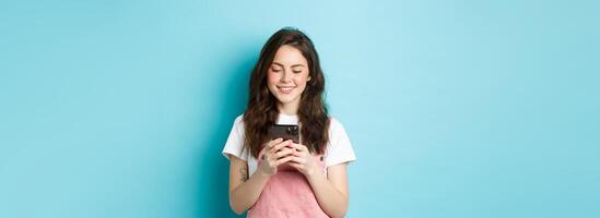 Portrait of young woman using smartphone, reading message or chatting in app, standing over blue background and smiling happy photo