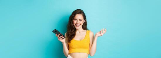 elegante contento mujer participación teléfono inteligente, riendo y sonriente seguro de sí mismo, chateando en móvil teléfono, en pie terminado azul antecedentes foto
