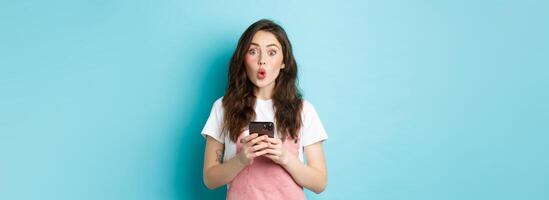 Portrait of brunette cute girl look surprised after using smartphone, holding mobile phone and say wow at camera with impressed face, blue background photo