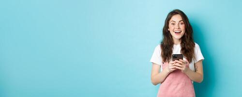 Portrait of excited brunette girl looking amazed at camera while using smartphone, holding mobile phone and smiling cheerful, standing against blue background photo