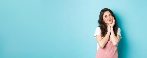 Portrait of beautiful girl dreaming, looking aside at logo banner and thinking, standing romantic against blue background, smiling and imaging something cute, blue background photo