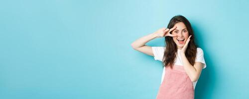 Portrait of excited attractive woman showing v-sign near eye, making kawaii gesture and smiling happy at camera, standing upbeat against blue background photo