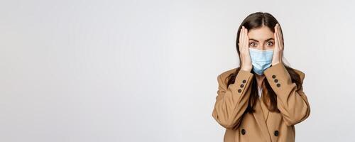 Covid-19 and people concept. Concerned, troubled office lady in medical face mask, holding hands on head and staring shocked at camera, white background photo