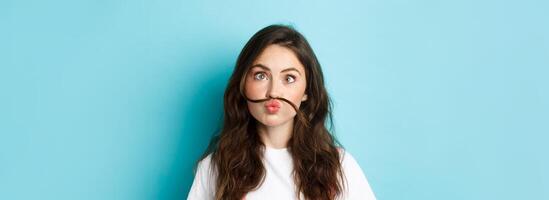 Close up portrait of funny silly girl playing, making squinting face and moustache with hair strand above lips, standing over blue background photo