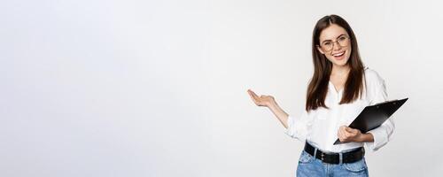 Smiling brunette woman, office lady showing way, pointing behind, demonstrating smth near her on copy space, standing over white background photo